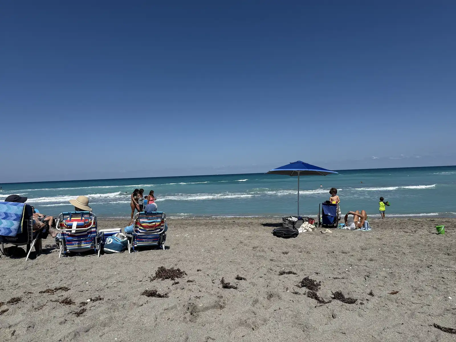 Beach chairs, coolers, bags and a blue umbrella sit on seaweed-dotted sand; several people sit and stand near the water with surf behind them under a blue sky.