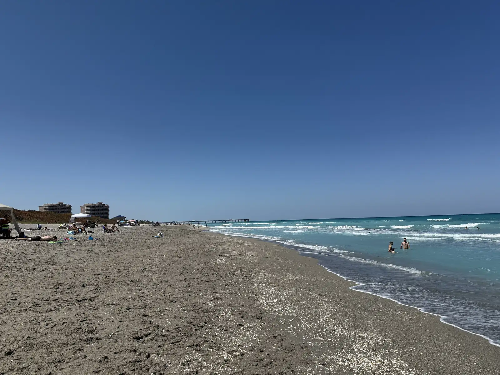 A broad tan beach curves north beside clear green-blue surf; swimmers in the shallows, beachgoers and umbrellas along the shore, two mid-rise buildings above the dunes and a long pier on the horizon, under a deep blue cloudless sky.