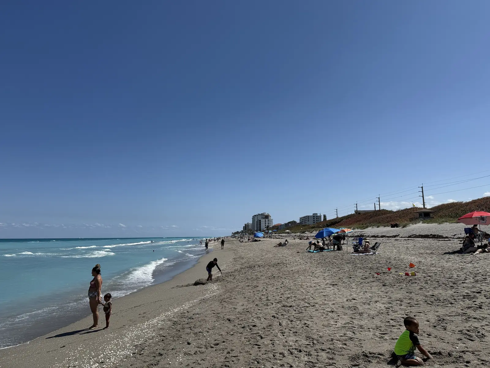 Looking south along a busy beach: a woman and a small child stand at the water's edge on the left, a boy plays in the sand in the foreground, umbrellas and beachgoers line the shore, and mid-rise buildings and utility poles rise above the dune.