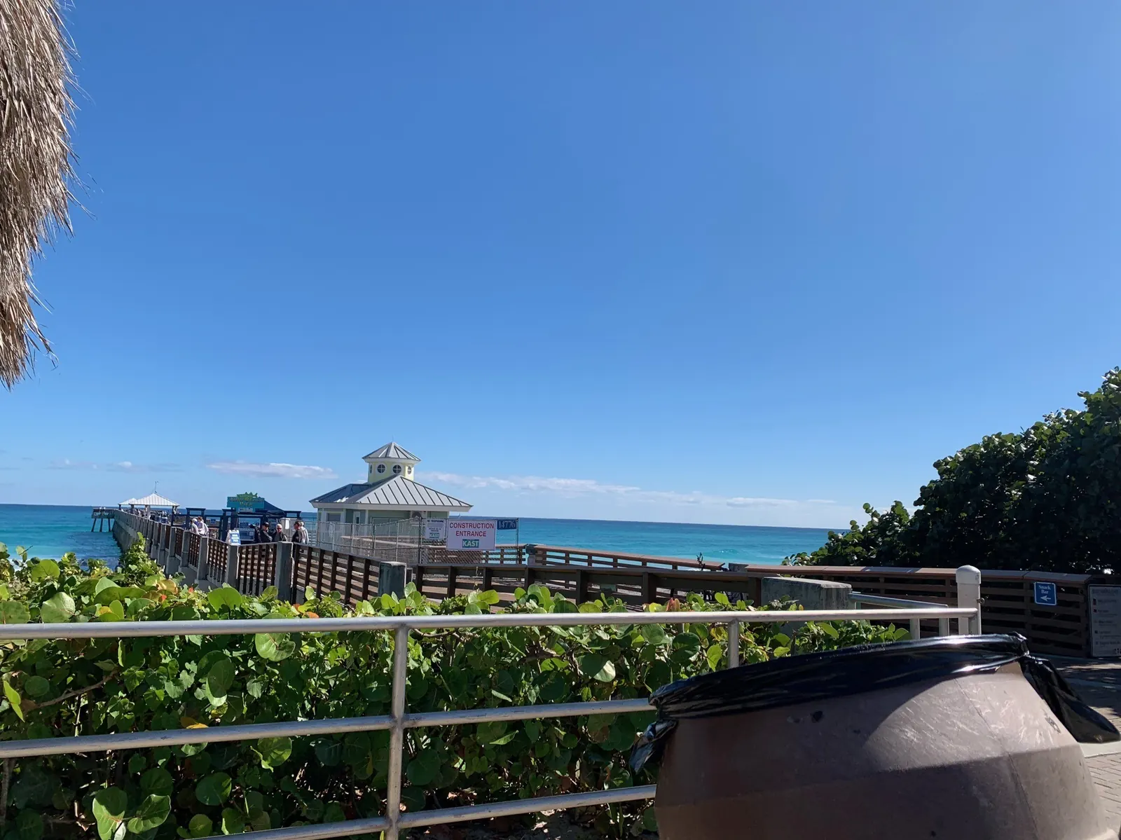 View past a metal railing and sea-grape shrubs toward the entrance of a wooden pier: a yellow-and-white cupola building, chain-link fencing and a sign reading 'CONSTRUCTION ENTRANCE' stand beside the walkway, a few people walk on the pier, and the turquoise ocean stretches to the horizon. A brown trash can fills the lower right.