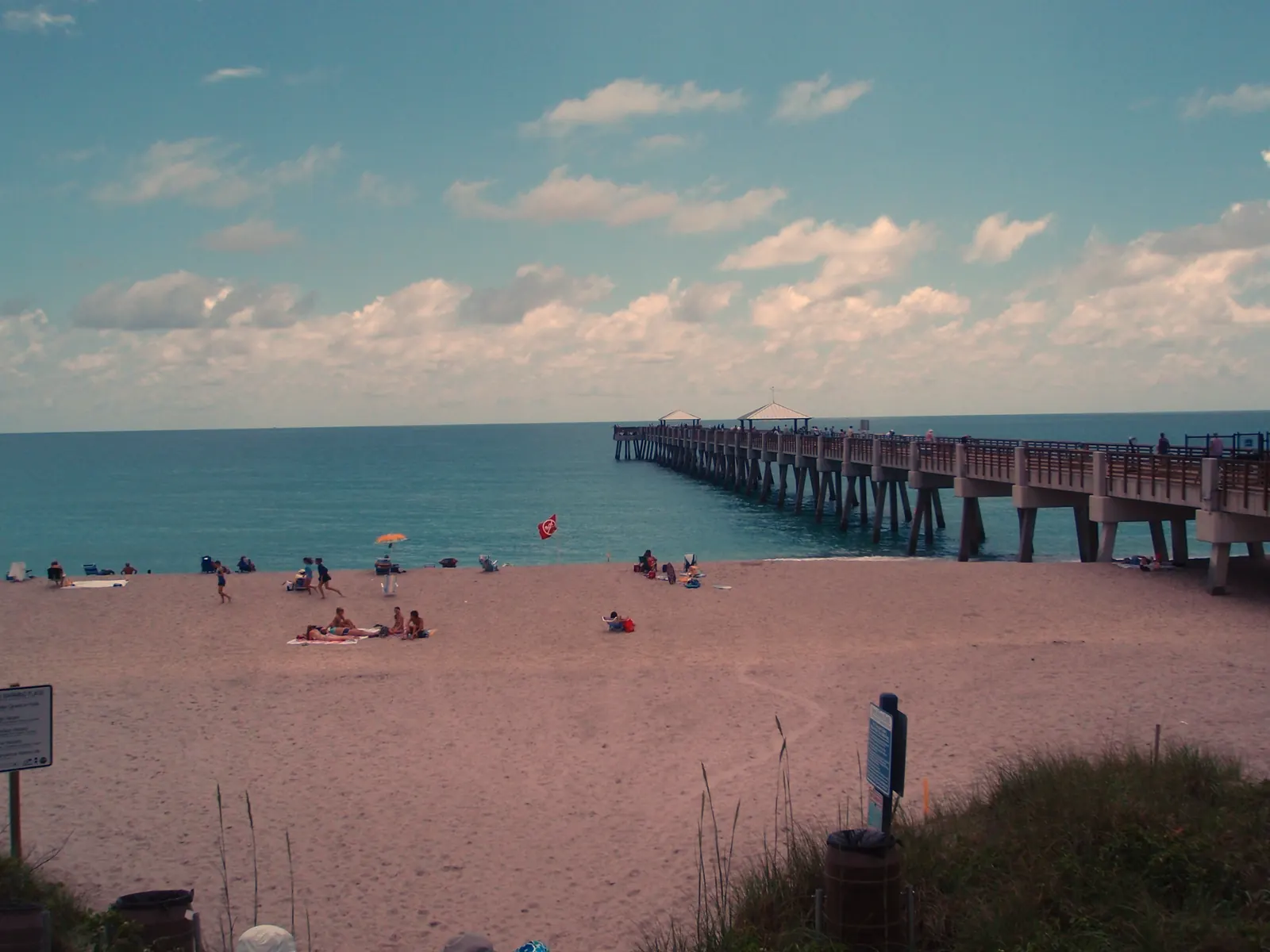 From a dune crossover with sea oats and a signpost, a wide sandy beach slopes down to blue water; sunbathers, a beach umbrella and a red flag are scattered on the sand, and a long pier with two roofed shelters runs out to sea on the right under puffy clouds.