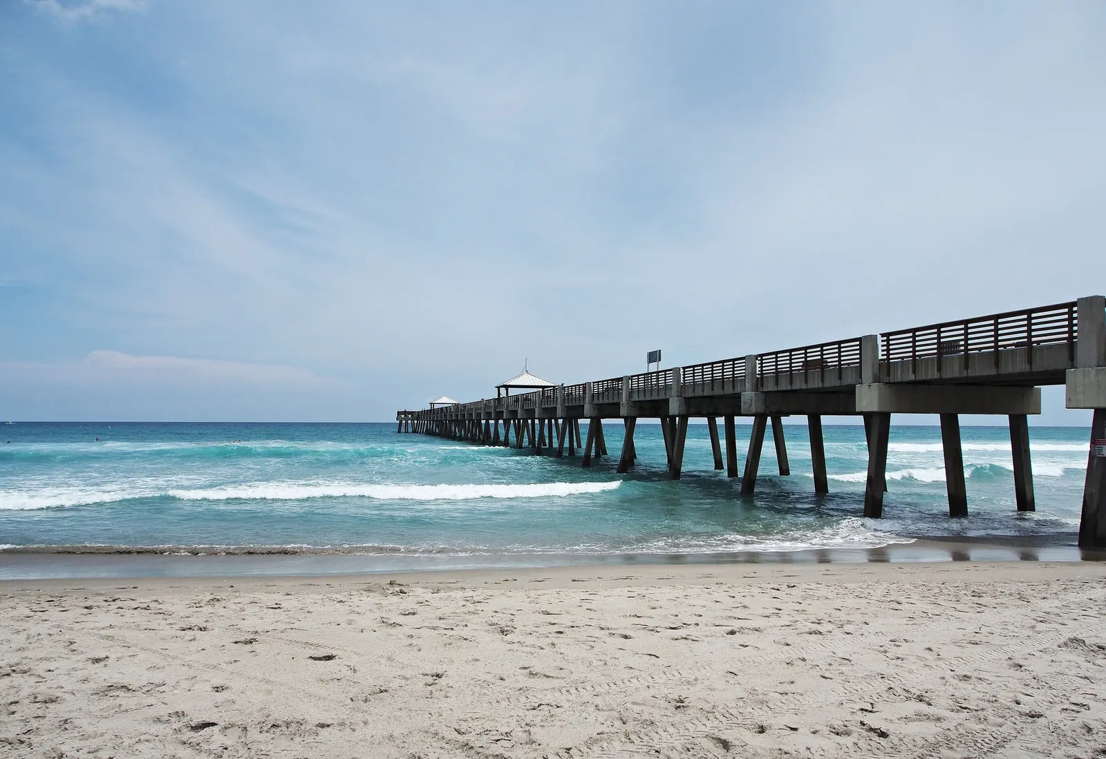 Low view from wet sand at the water's edge looking along a long concrete fishing pier on square pilings that runs out into turquoise surf; two white-roofed shelters stand near the far end. Small breaking waves, a wide beach of footprinted sand in the foreground, and a pale overcast sky.