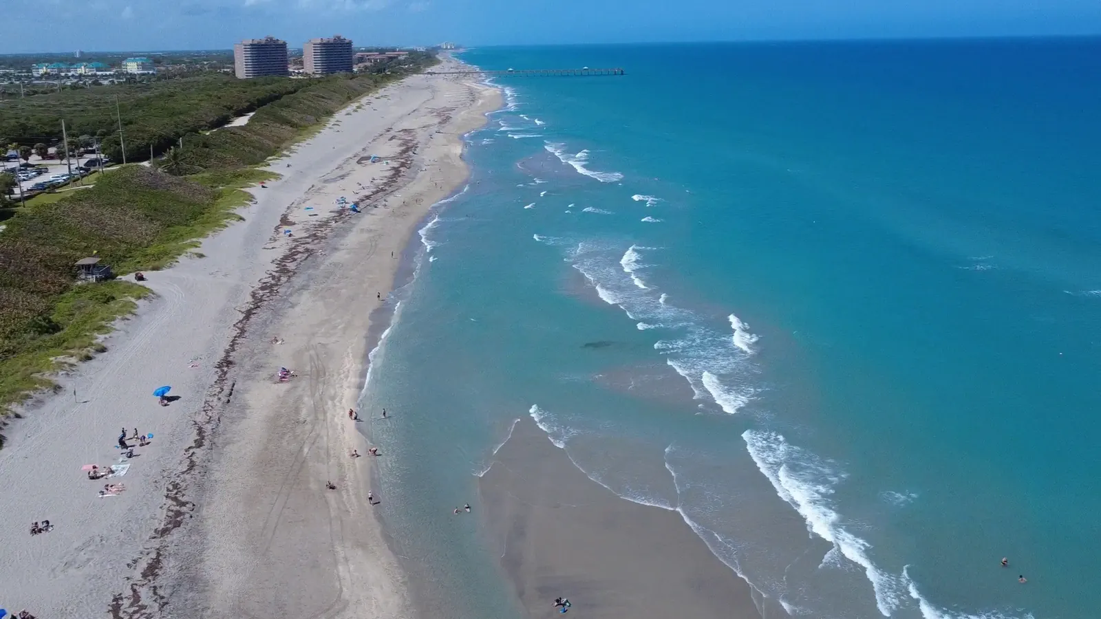 Aerial view looking north along a long, narrow sand beach: turquoise water and white surf on the right, green dune vegetation and a parking lot on the left, scattered beachgoers and umbrellas, two tall towers in the distance and a thin pier reaching out to sea at the horizon.