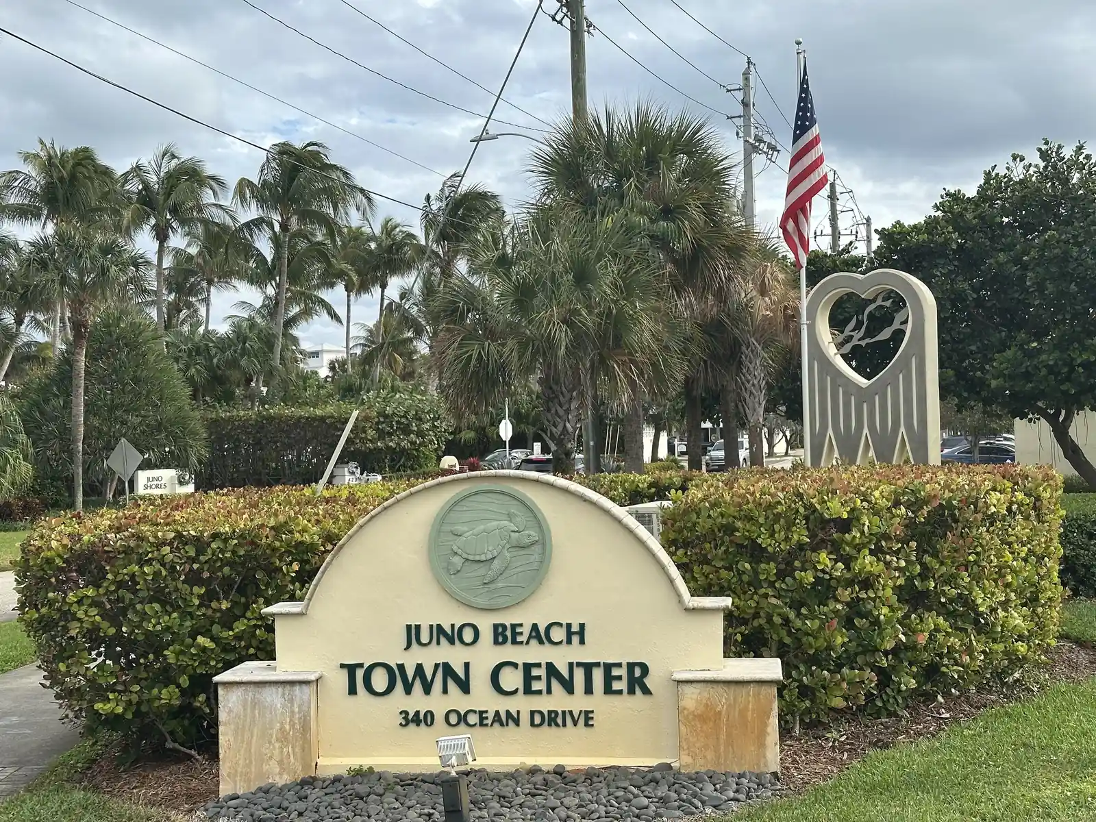 A cream monument sign with a sea-turtle medallion reads 'Juno Beach Town Center, 340 Ocean Drive', set in clipped hedges; behind it are palms, an American flag and a tall heart-shaped sculpture, under a cloudy sky with power lines.