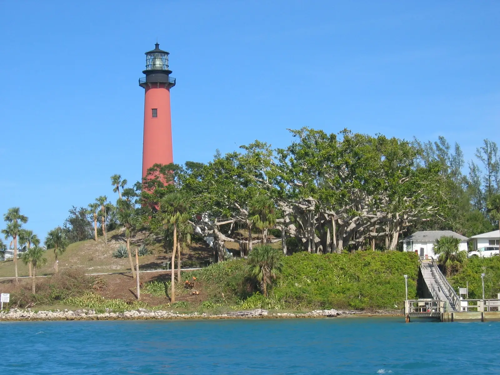 A tall red lighthouse with a black lantern stands on a green, tree-covered hill above blue water; a stairway leads down to a small dock at right and white buildings sit among banyan-like trees.