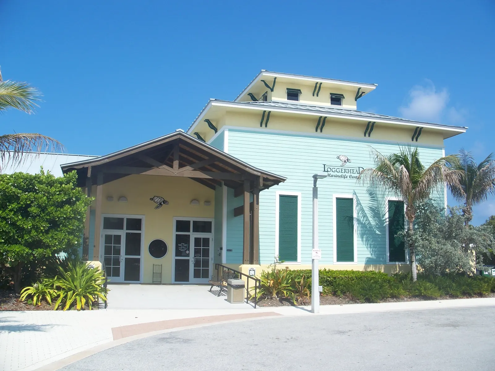 Front entrance of a pale aqua two-storey building with yellow trim, green shutters and a timber-framed porch; a turtle emblem is above the door and lettering on the wall reads 'Loggerhead Marinelife Center', with palms and tropical plants beside a curved drive.