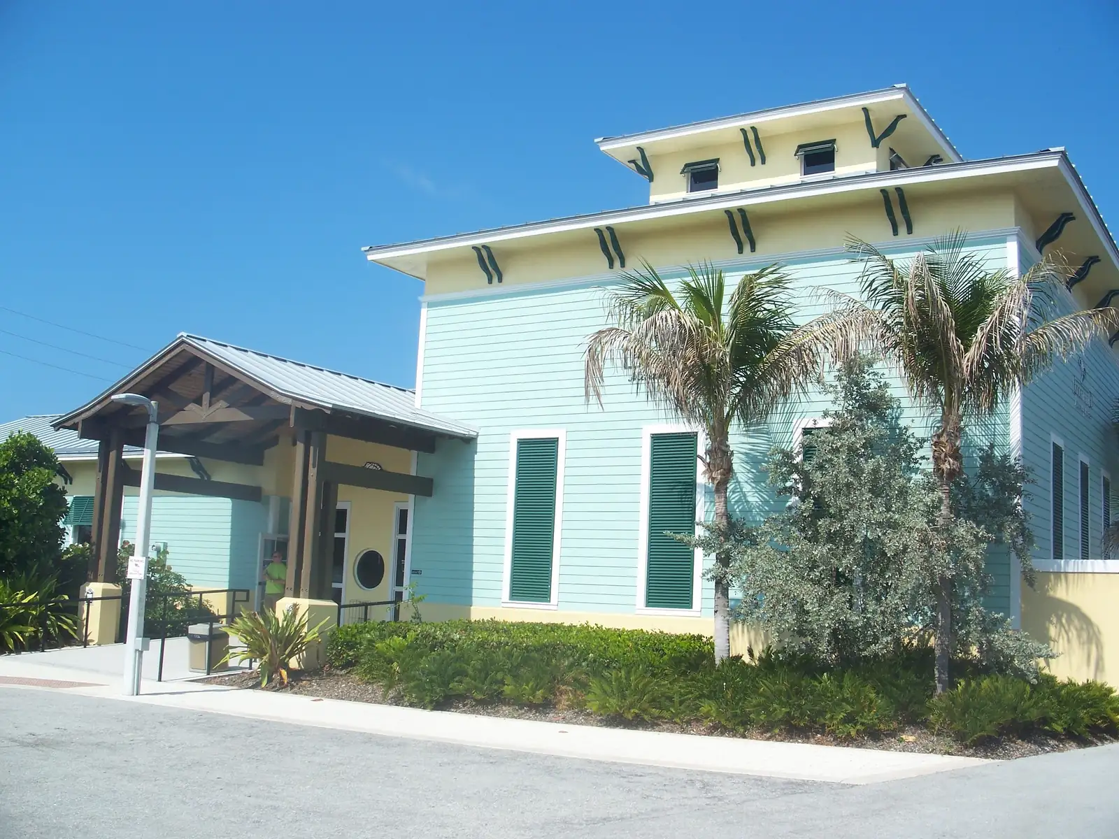 Corner view of a pale aqua building with a yellow upper storey, green shutters and green decorative brackets, a timber porch at left, and palms and silver-leaved shrubs in front, under a blue sky.